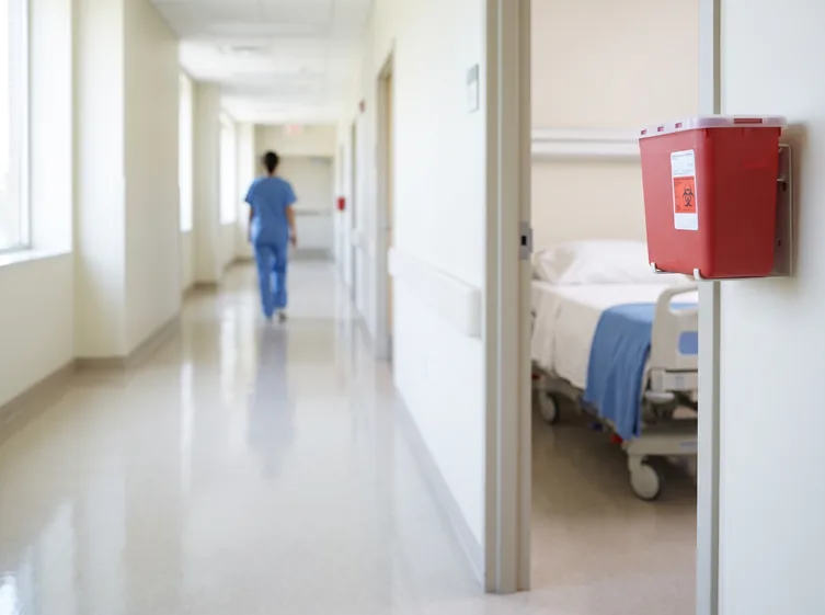 A red sharps container mounted on the wall outside a hospital patient room, with a nurse walking away down the corridor.