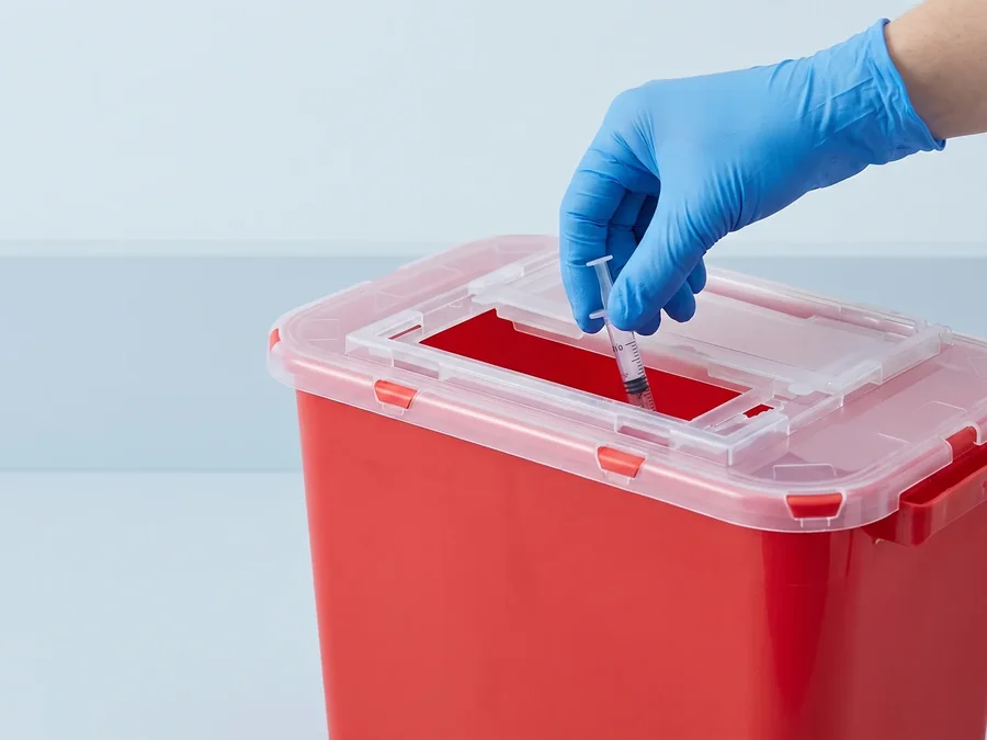 A gloved hand dropping a used syringe through the lid of a red rigid sharps container.